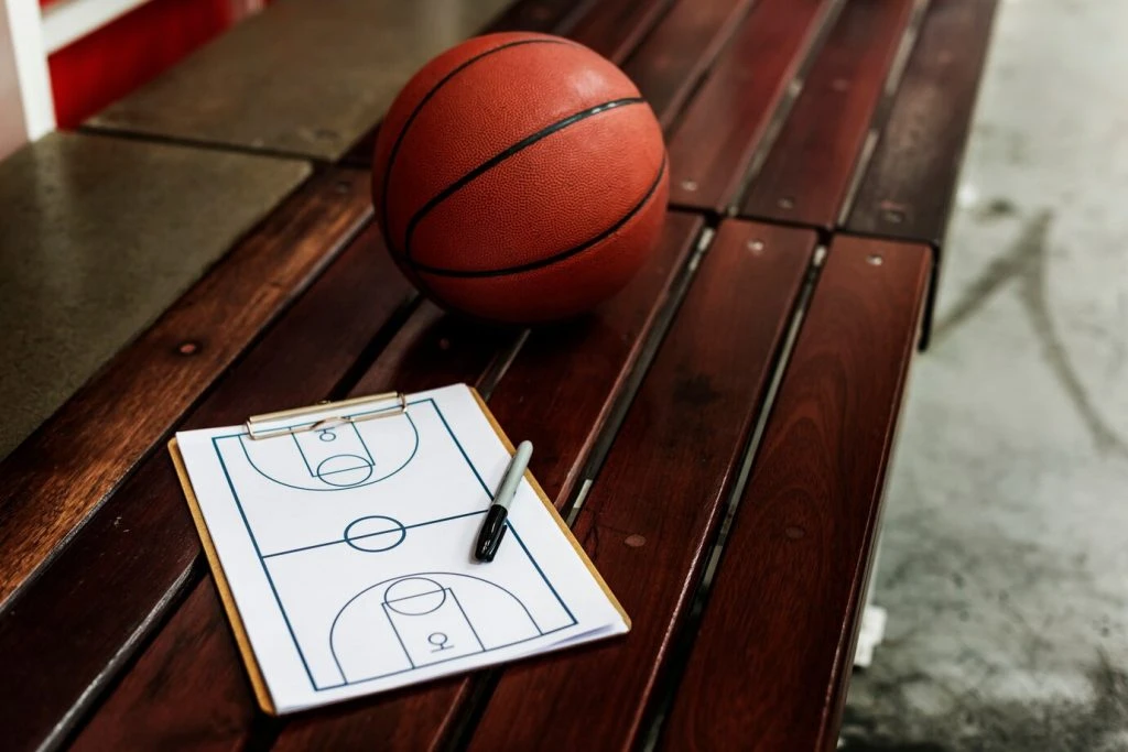 Pelota de basquetbol apoyada en una banca de madera junto a una planilla y un marcador con el esquema de un campo de baloncesto para planear estrategias de juego. 