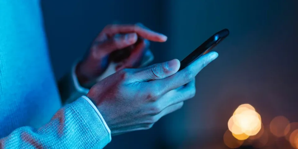 Manos de un hombre joven interactuando con un teléfono inteligente bajo una luz azul