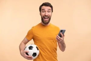 Hombre feliz con barba mirando a cámara con un teléfono inteligente en una mano y una pelota de fútbol en la otra 