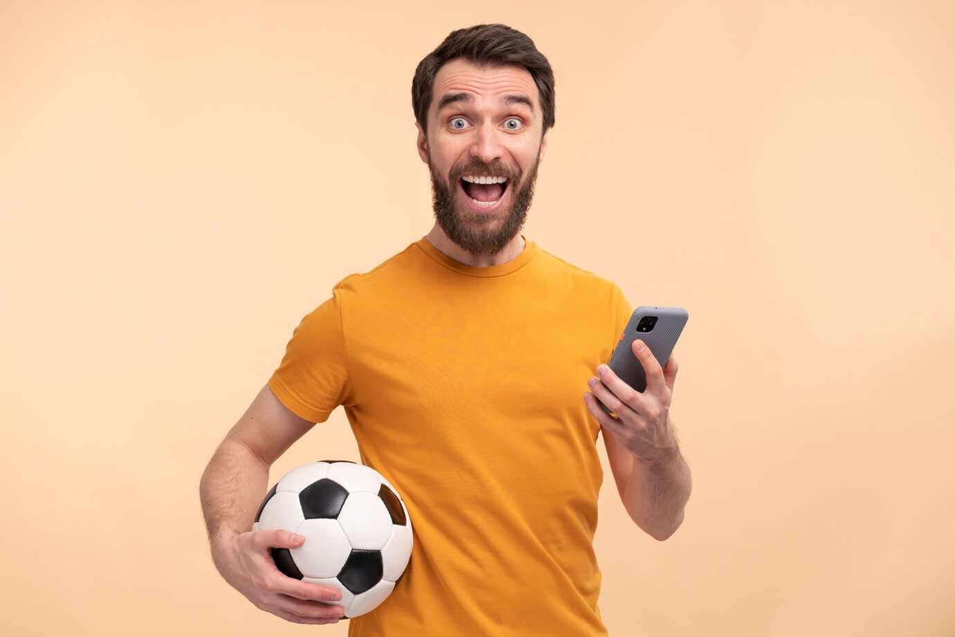 Hombre feliz con barba mirando a cámara con un teléfono inteligente en una mano y una pelota de fútbol en la otra