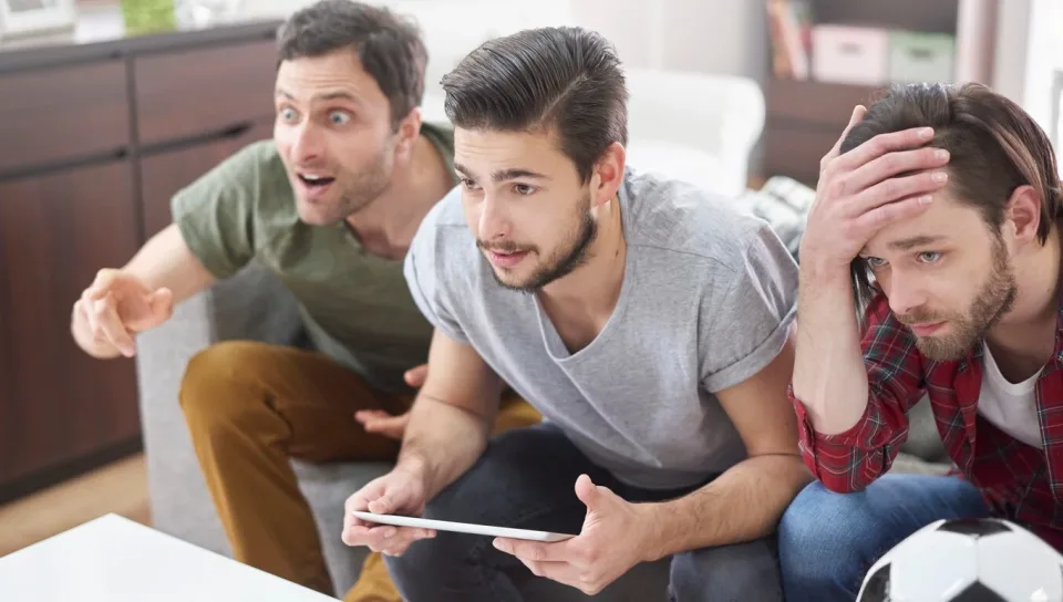 Tres hombres jóvenes viviendo apasionadamente un evento por televisión desde el sofá de su casa